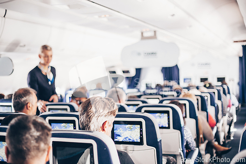 Image of Interior of airplane with passengers on seats and stewardess in uniform walking the aisle, serving people. Commercial economy flight service concept.