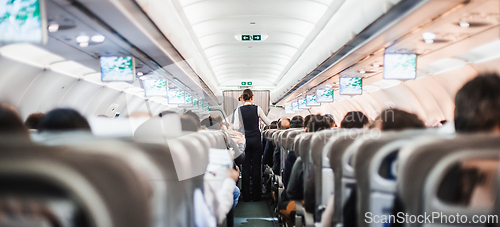 Image of Interior of airplane with passengers on seats and stewardess in uniform walking the aisle, serving people. Commercial economy flight service concept.