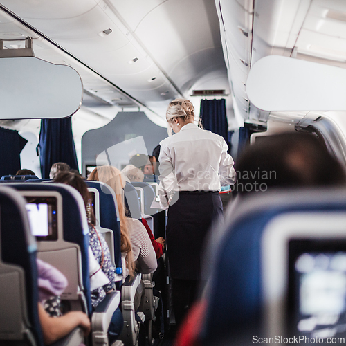 Image of Interior of airplane with passengers on seats and stewardess in uniform walking the aisle, serving people. Commercial economy flight service concept.