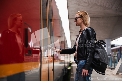 Image of Young blond woman in jeans, shirt and leather jacket wearing bag and sunglass, presses door button of modern speed train to embark on train station platform. Travel and transportation.