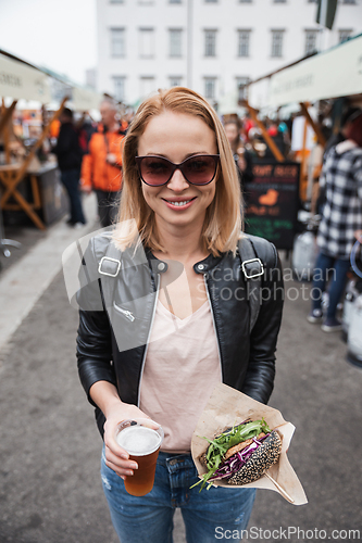 Image of Beautiful young woman holding delicious organic salmon vegetarian burger and homebrewed IPA beer on open air beer an burger urban street food festival in Ljubljana, Slovenia.