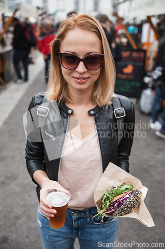 Image of Beautiful young woman holding delicious organic salmon vegetarian burger and homebrewed IPA beer on open air beer an burger urban street food festival in Ljubljana, Slovenia.