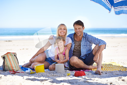 Image of Parents, girl and sand castle in portrait by ocean, blanket and umbrella with hug on holiday in summer. Father, mother and daughter with picnic at beach for vacation in nature with love in sunshine