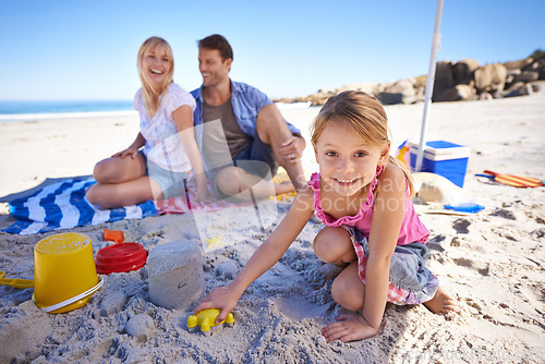 Image of Parents, girl and sandcastle in portrait by sea, blanket and excited with building for holiday in summer. Father, mother and daughter with family picnic by ocean for vacation with smile in sunshine