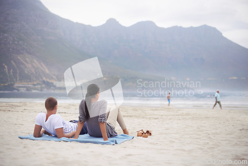 Image of Couple, beach and vacation on blanket, picnic or relaxing on seashore by mountains. Romantic, honeymoon and love between happy husband and wife, married and date on ocean coast enjoying sunshine