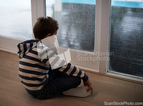 Image of Rain, window and sad boy child on a floor unhappy, upset or annoyed by bad weather in his home. Glass, depression and kid in a living room frustrated by storm, cold or unexpected winter disaster