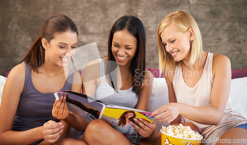 Image of Friends, smile and magazine with popcorn on bed for reading, discussion and girls night in home with pyjamas. Women, excited and happiness with paper in house for bonding, conversation and snack
