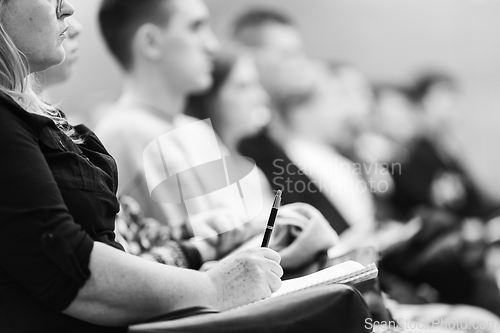 Image of Female hands holding pen and notebook, making notes at conference lecture. Event participants in conference hall. Black and white image.