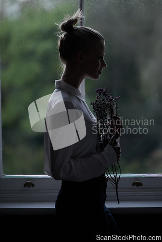 Image of Woman, flowers and window for in home, dark room and mysterious aesthetic for person or artistic pose. Female artist, dim light and silhouette with thinking, fashion and morning setting in dawn