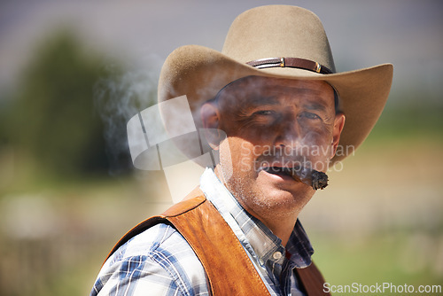 Image of Portrait, cowboy and man smoking cigar at farm in the rural countryside in Texas. Ranch, face and serious person with tobacco in western hat outdoor, nicotine and casual clothes in nature at stable
