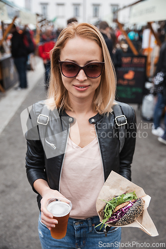 Image of Beautiful young woman holding delicious organic salmon vegetarian burger and homebrewed IPA beer on open air beer an burger urban street food festival in Ljubljana, Slovenia.