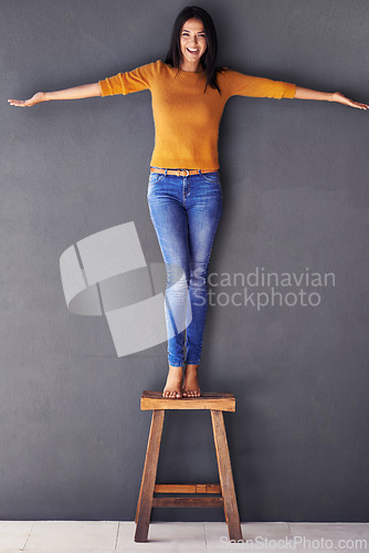 Image of Woman, confident and stand on chair in portrait, smile and fashion on wall background. Female person, clothes and happy lady on stool, apparel and trends in living room at home in house
