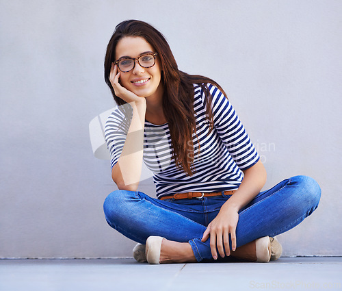 Image of Happy, fashion and portrait of woman on floor with wall background for casual or trendy style. Smile, model and relax with confident young person legs crossed in glasses for casual clothing outfit