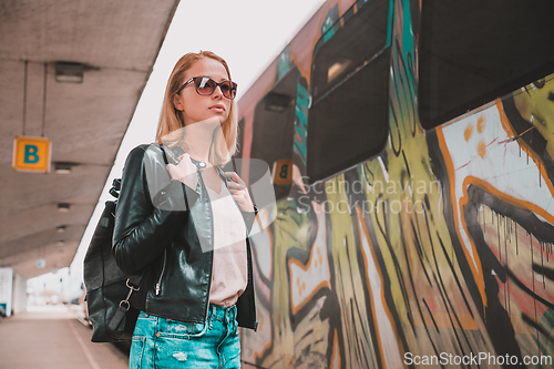 Image of Young blond woman in jeans, shirt and leather jacket wearing bag and sunglass, embarking modern speed train on train station platform. Travel and transportation.