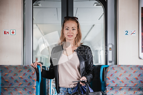 Image of Young blond woman in jeans, shirt and leather jacket holding her smart phone and purse while riding modern speed train arriving to final train station stop. Travel and transportation.