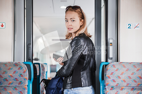 Image of Young blond woman in jeans, shirt and leather jacket holding her smart phone and purse while riding modern speed train arriving to final train station stop. Travel and transportation.