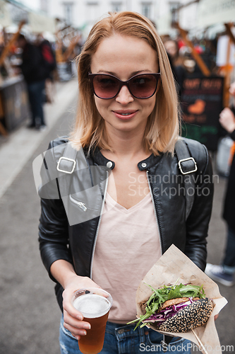 Image of Beautiful young woman holding delicious organic salmon vegetarian burger and homebrewed IPA beer on open air beer an burger urban street food festival in Ljubljana, Slovenia.
