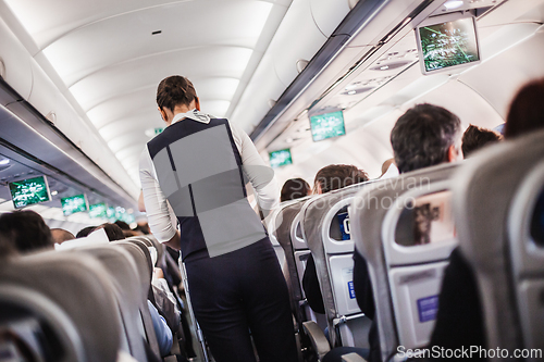 Image of Interior of airplane with passengers on seats and stewardess in uniform walking the aisle, serving people. Commercial economy flight service concept.