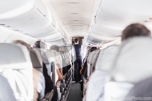 Image of Interior of airplane with passengers on seats and stewardess in uniform walking the aisle, serving people. Commercial economy flight service concept.