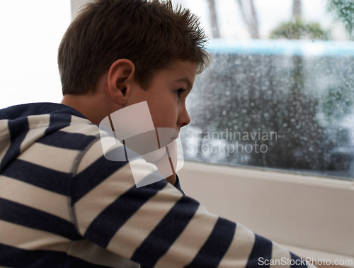 Image of Window, rain and sad boy child on a floor unhappy, upset or annoyed by bad weather in his home. Glass, depression and kid in a living room frustrated by storm, cold or unexpected winter disaster