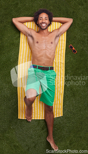 Image of Happy man, portrait and lying with towel on green grass above for relaxation or outdoor summer holiday. Top view of young male person with smile in relax for swimwear or fashion on field in nature