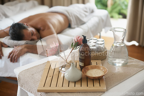 Image of Salt, flowers and man in spa to relax on bed or break with luxury pamper treatment tools on table. Protea, facial oil or person at resort and salon for natural healing benefits or massage with water