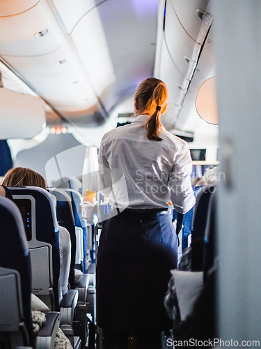 Image of Interior of airplane with passengers on seats and stewardess in uniform walking the aisle, serving people. Commercial economy flight service concept.