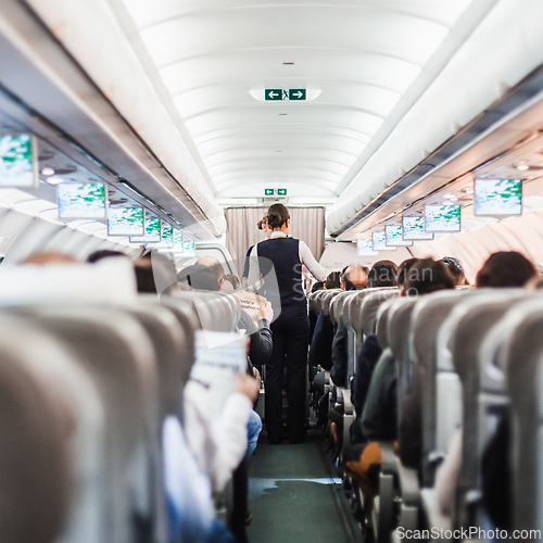 Image of Interior of airplane with passengers on seats and stewardess in uniform walking the aisle, serving people. Commercial economy flight service concept.