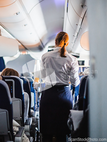 Image of Interior of airplane with passengers on seats and stewardess in uniform walking the aisle, serving people. Commercial economy flight service concept.