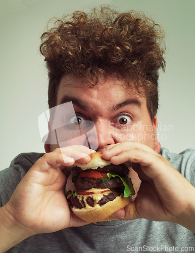 Image of Takeaway, burger and food for man in studio with meal for big eater, plus size or greed. Young person with meat in mouth for hunger, silly and bite indoor for delicious, taste and sauce for portrait