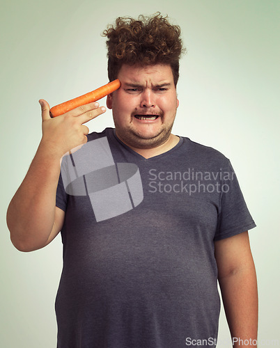Image of Depression, carrot and hand gun with plus size man in studio on gray background for diet, health or nutrition. Sad, unhappy and vegetable to head of young person in conflict with vegetarian food