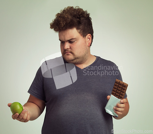 Image of Man, apple and chocolate in studio with choice for diet, healthy food and nutrition for wellness. Male person, plus size and thinking with decision for fruit, sweet dessert and snack for lifestyle