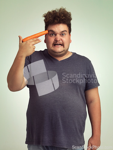 Image of Portrait, vegetable and hand gun with plus size man in studio on gray background for diet, health or nutrition. Anger, depression and carrot to head of young person in conflict with vegetarian food