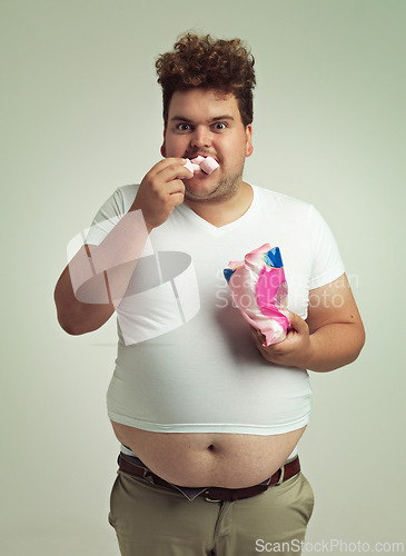 Image of Portrait, hunger and marshmallow with plus size man in studio on gray background for unhealthy eating. Food, hungry for sweets and candy with young person with snack bag or packet for greed or belly
