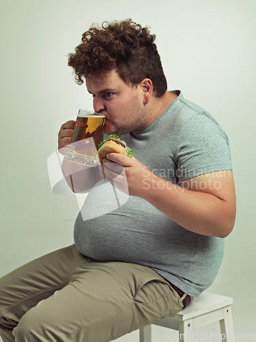 Image of Man, drink and beer with burger in studio for unhealthy diet, fast food and nutrition on chair. Plus size, hungry and male person with alcohol on stool for dinner, meal and beverage in glass