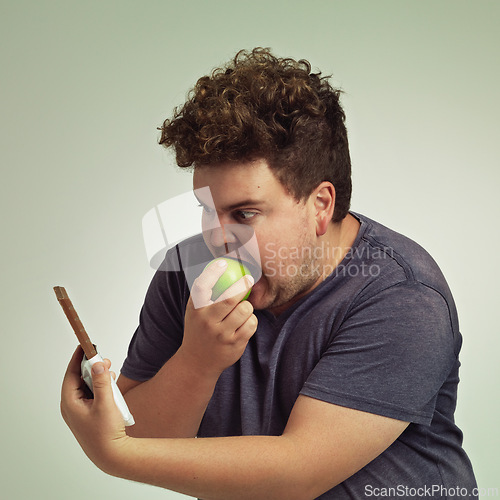 Image of Guy, apple and chocolate in studio with choice of junk food, sweet or diet for wellness. Plus size, male person eating and decision, fruit or candy for nutrition, lose weight and healthy lifestyle