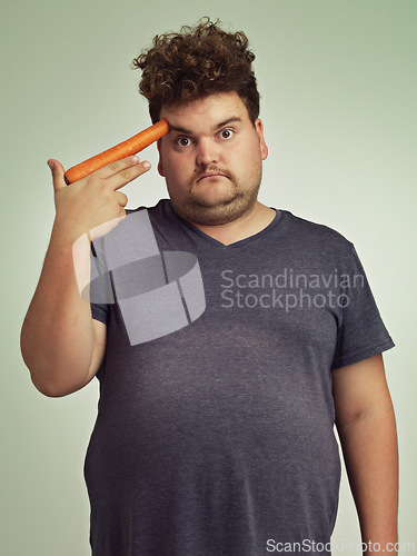 Image of Portrait, carrot and hand gun with plus size man in studio on gray background for diet, health or nutrition. Depression, sad and vegetable to head of young person in conflict with vegetarian food