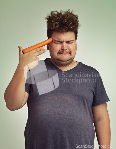 Image of Depression, vegetable and hand gun with plus size man in studio on gray background for diet, health or nutrition. Sad, unhappy and carrot to head of young person in conflict with vegetarian food