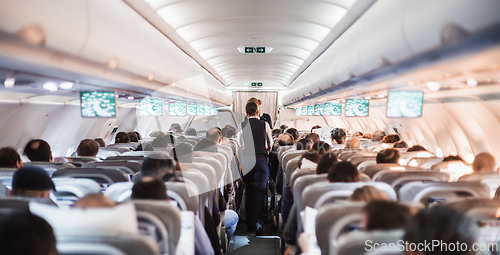 Image of Interior of airplane with passengers on seats and stewardess in uniform walking the aisle, serving people. Commercial economy flight service concept.