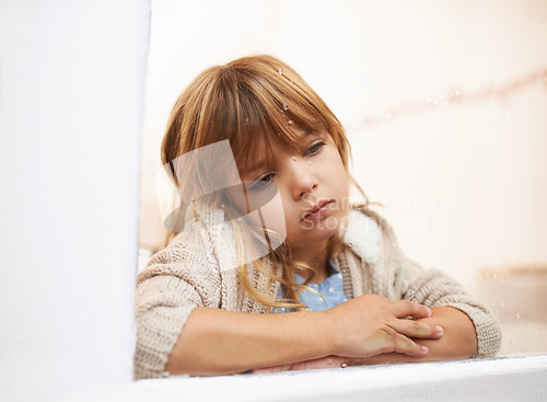 Image of Window, sitting and child in house for bored, lonely or disappointed for rain or weather outdoor. Young girl or kid and sadness or sick in home for cold season and wait for play for childhood