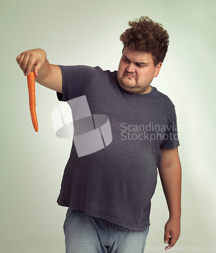 Image of Plus size, diet and disgusted man with carrot for health, nutrition and wellness in studio by white background. Weight loss, vegetable and unhappy male person for detox, wellbeing and vegan food