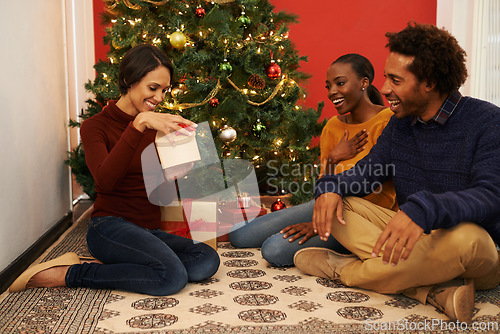 Image of Christmas, tree and group of friends with present and opening surprise on festive holiday or vacation with happiness. Excited, people and giving gift to person in home lounge with love and kindness