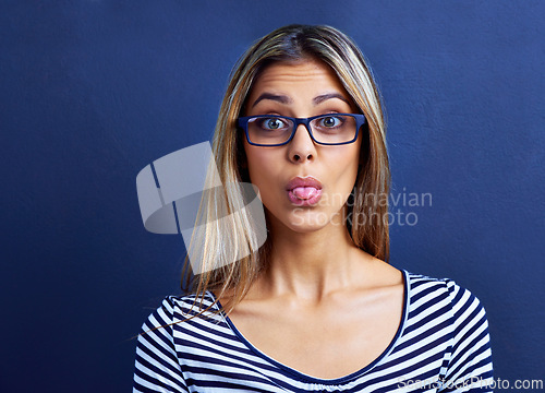 Image of Funny, face and woman with glasses in studio for tongue out, silly or humor on blue background. Need, portrait and female model with emoji expression, gesture or geek, aesthetic or quirky personality