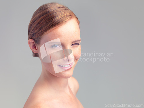 Image of Portrait, wink and skincare of happy woman for beauty, shine or mockup space in studio isolated on a gray background. Face, smile and ginger model with cosmetics, makeup or treatment for dermatology