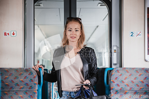 Image of Young blond woman in jeans, shirt and leather jacket holding her smart phone and purse while riding modern speed train arriving to final train station stop. Travel and transportation.