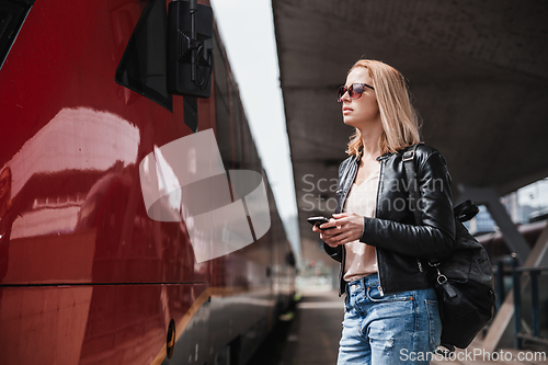 Image of Young blond woman in jeans, shirt and leather jacket wearing bag and sunglass, embarking red modern speed train on train station platform. Travel and transportation.