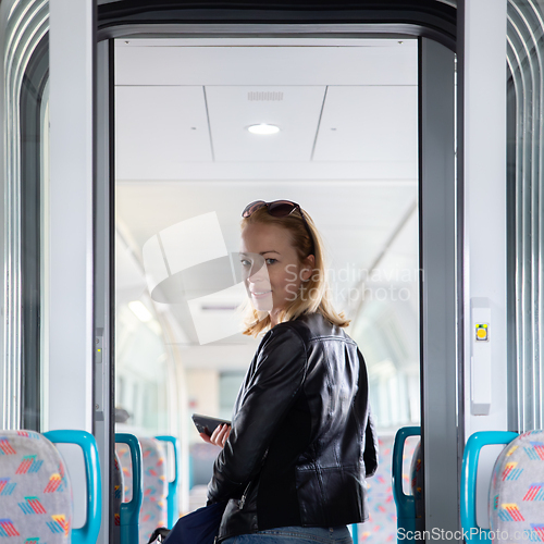Image of Young blond woman in jeans, shirt and leather jacket holding her smart phone and purse while riding modern speed train arriving to final train station stop. Travel and transportation.