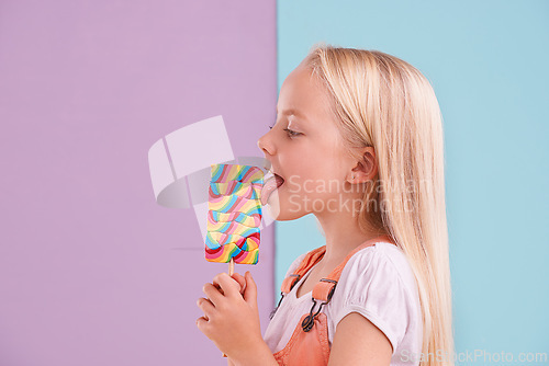 Image of Studio, girl and lick with lollipop, dessert and choice for snack and childhood. Child, sweets and yummy candy for tasty, eating and happy or excited on vibrant split pastel pink and blue background