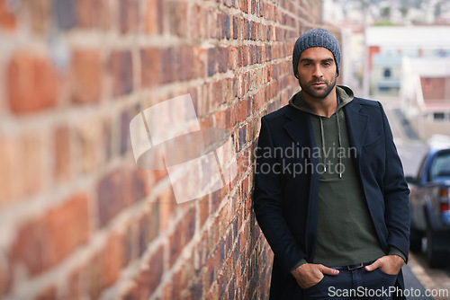 Image of Portrait, confidence and man on brick wall in city in casual clothes, outfit or fashion for winter outdoor. Serious, cool and stylish person in urban town in beanie, hoodie and trendy jacket in Spain