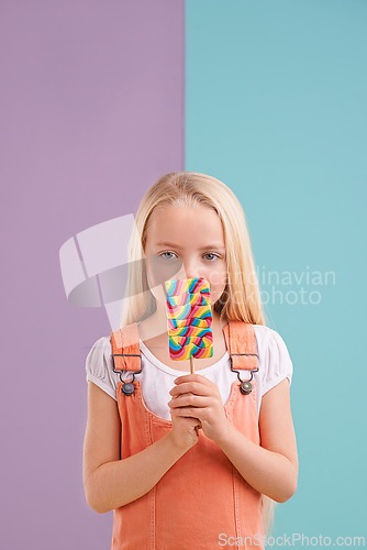 Image of Studio, child and hiding with lollipop, desert and playful for snack and childhood. Girl, sweets and yummy candy for tasty, eating and happiness on vibrant split pastel pink and blue background